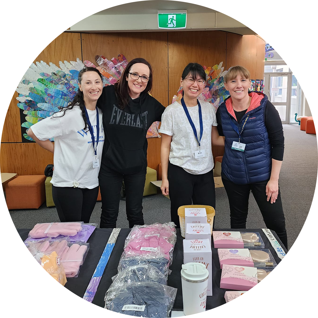 Four volunteers standing behind a school Mother's Day stall, smiling at the camera. The table displays various gifts for sale, including pink gloves, packaged clothing, and boxes. A colourful, feather-like mural is visible on the wall behind them.