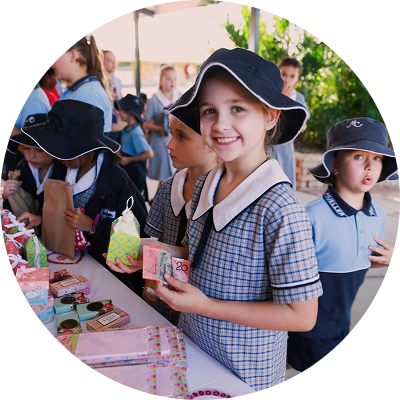 A student in a school uniform and hat stands at a Mother's Day stall, holding a twenty-dollar note and smiling at the camera. The stall table is covered with various colourful gift items.