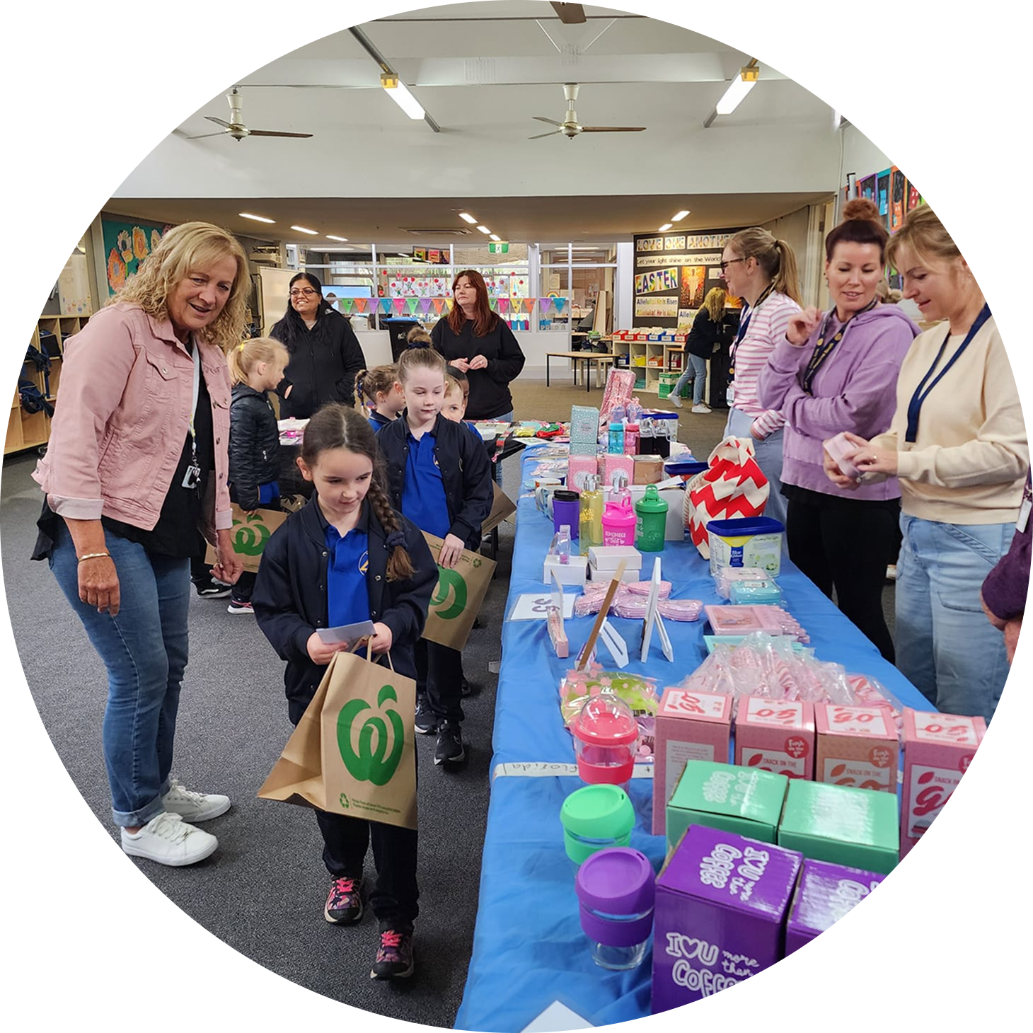 A busy Mother's Day fundraising stall at a school, featuring a table covered in blue cloth with various gifts for sale, including mugs, drink bottles, and packaged items. Several adults and children in school uniform are gathered around the table, with one child holding a reusable shopping bag.