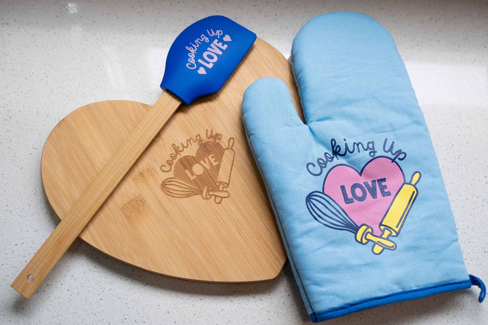 A Mother's Day fundraising set featuring a heart-shaped wooden cutting board, a blue "Cooking up Love" spatula, and a matching blue oven mitt.